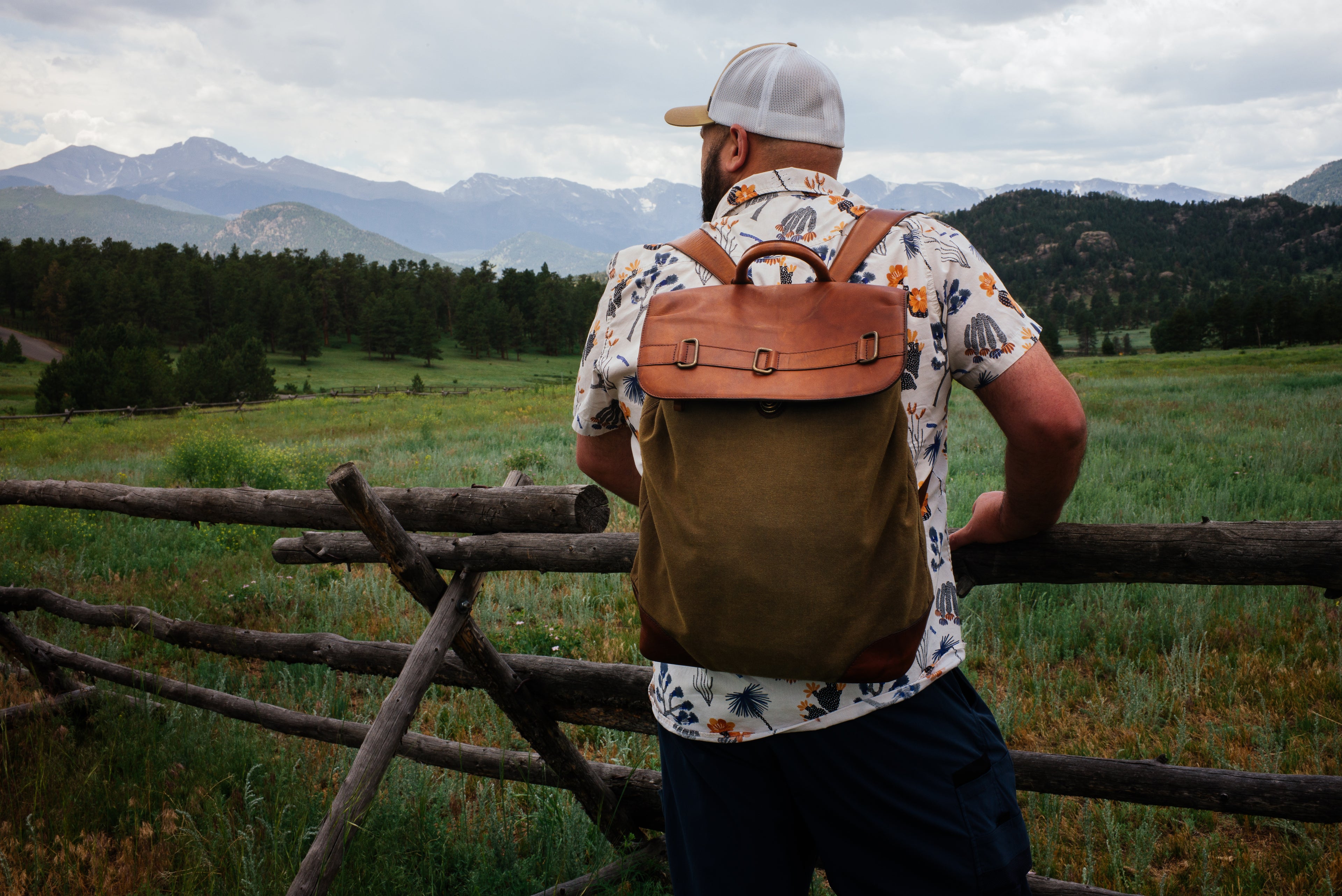 Man with a leather backpack standing by a wooden fence in a scenic landscape with mountains.